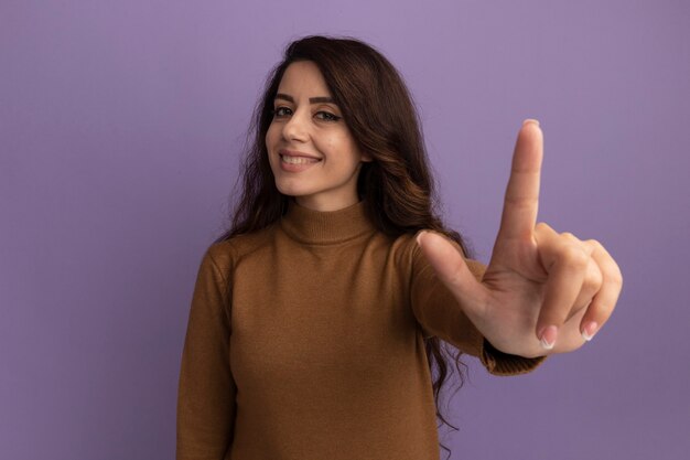 Smiling young beautiful girl wearing brown turtleneck sweater points at front isolated on purple wall