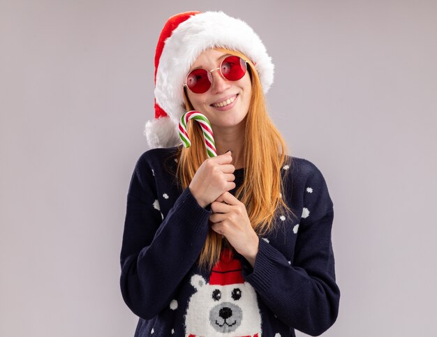 クリスマスの帽子と白い壁に隔離された顔の周りにクリスマス キャンディーを保持しているメガネを身に着けている笑顔の美しい少女