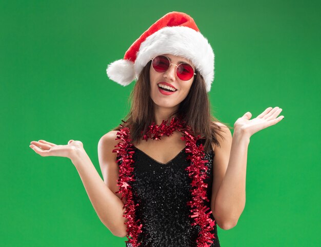 Smiling young beautiful girl wearing christmas hat and glasses with garland on neck spreading hands isolated on green wall