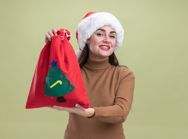 올리브 녹색 배경에 고립 된 카메라에서 크리스마스 가방을 들고 크리스마스 모자를 쓰고 웃는 젊은 아름 다운 소녀