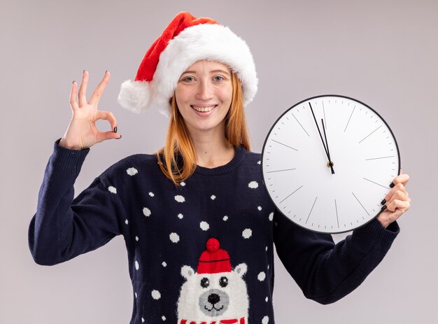 Улыбающаяся молодая красивая девушка в новогодней шапке держит настенные часы, показывая хороший жест, изолированные на белой стене