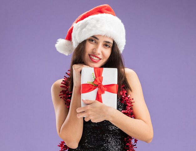 Smiling young beautiful girl wearing christmas hat with garland on neck holding gift box isolated on purple background