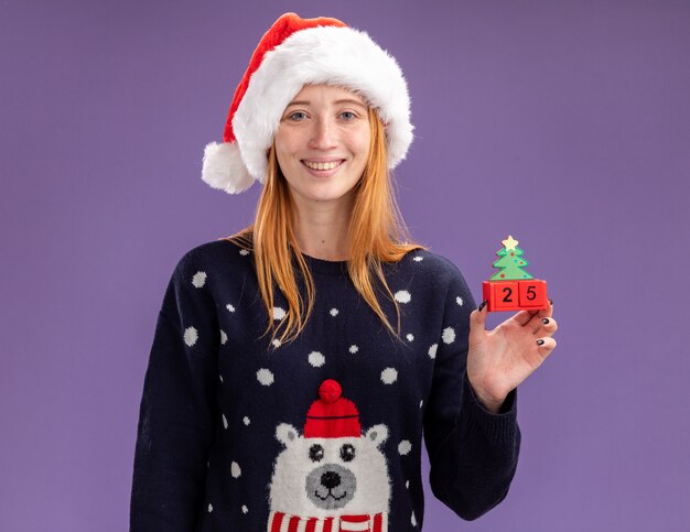 보라색 배경에 고립 된 크리스마스 장난감을 들고 안경 크리스마스 스웨터와 모자를 입고 젊은 아름 다운 소녀 미소