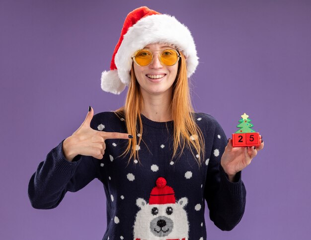 Smiling young beautiful girl wearing christmas sweater and hat with glasses holding and points at christmas toy isolated on purple background
