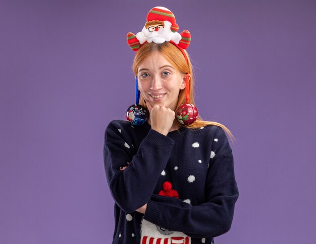 Sorridente giovane bella ragazza che indossa un maglione di natale con cerchietto per capelli di natale appeso palle di natale sull'orecchio mettendo la mano sotto il mento isolato sul muro viola