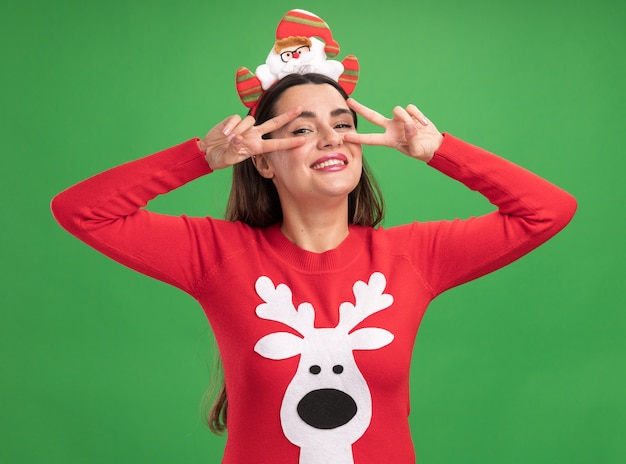 Smiling young beautiful girl wearing christmas sweater with christmas hair hoop showing peace gesture isolated on green wall