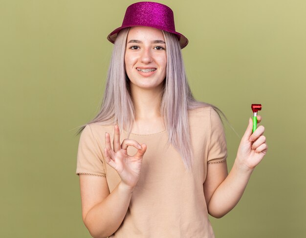 Smiling young beautiful girl wearing dental braces with party hat holding party whistle showing okay gesture 