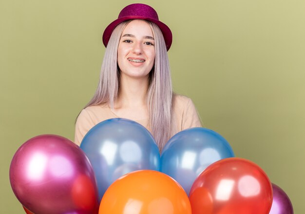 Smiling young beautiful girl wearing dental braces with party hat standing behind balloons 