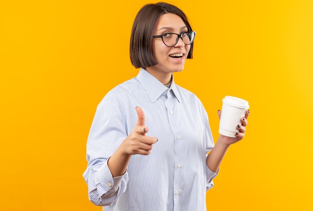 Smiling young beautiful girl wearing glasses holding cup of coffee points at camera 