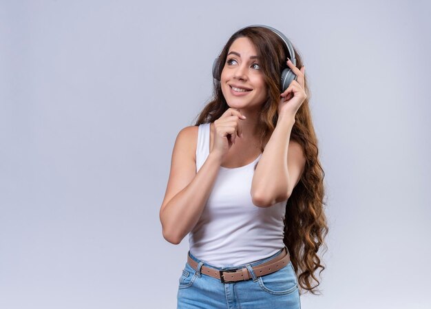 Smiling young beautiful girl wearing headphones listening to music looking at left side with one hand on headphone and another one on chin  with copy space