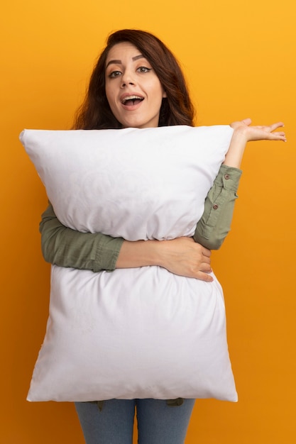 Smiling young beautiful girl wearing olive green t-shirt hugged pillow spreading hand isolated on yellow wall