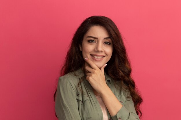 Smiling young beautiful girl wearing olive green t-shirt putting hand on chin isolated on pink wall