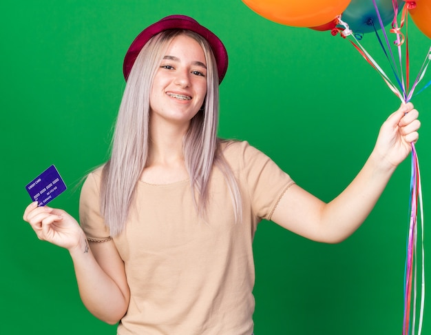 Улыбающаяся молодая красивая девушка в шляпе и подтяжках держит воздушные шары с кредитной картой