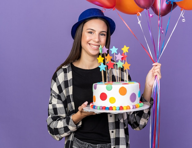 Smiling young beautiful girl wearing party hat holding balloons with cake isolated on blue wall