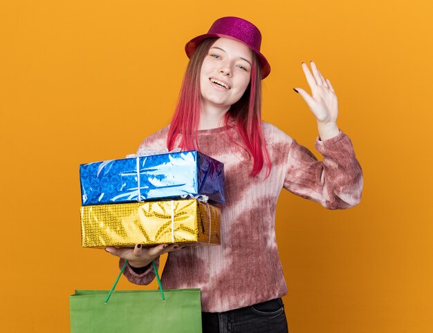 Улыбающаяся молодая красивая девушка в партийной шляпе держит подарочный пакет с подарочными коробками, показывая приветственный жест, изолированный на оранжевой стене
