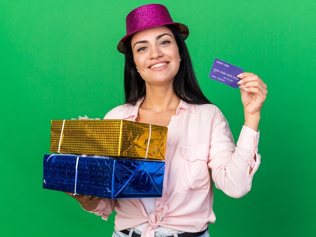 Улыбающаяся молодая красивая девушка в партийной шляпе держит подарочные коробки с кредитной картой, изолированной на зеленой стене