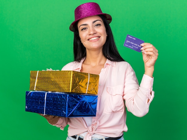 Smiling young beautiful girl wearing party hat holding gift boxes with credit card isolated on green wall