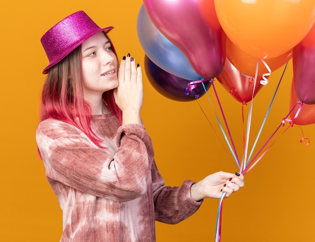 Smiling young beautiful girl wearing party hat holding and looking at balloons calling someone 