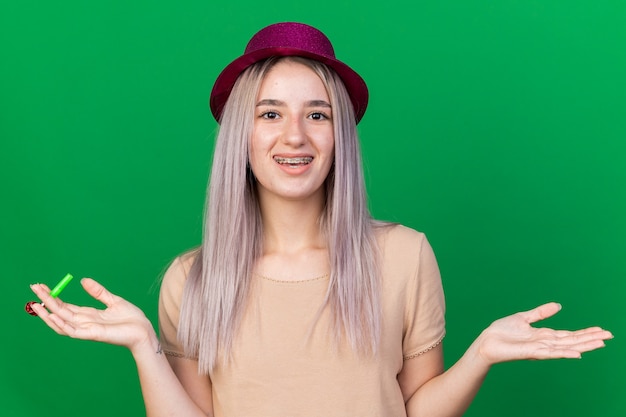 Улыбающаяся молодая красивая девушка в партийной шляпе, держащая партийный свисток, разводит руками, изолированными на зеленой стене