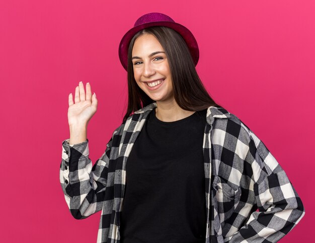 Smiling young beautiful girl wearing party hat points with hand at behind isolated on pink wall with copy space