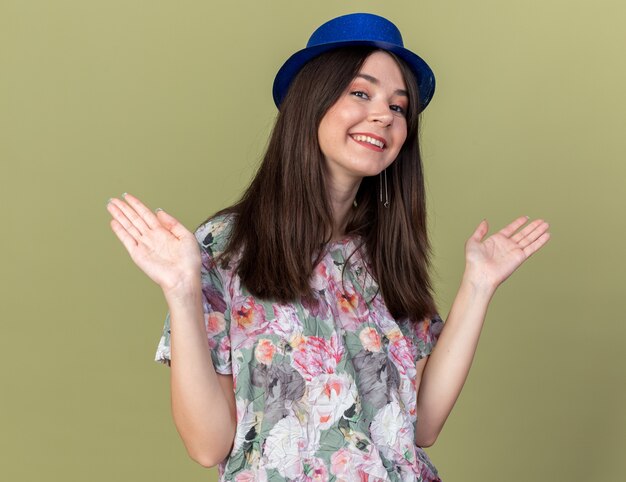 Smiling young beautiful girl wearing party hat spreading hands isolated on olive green wall