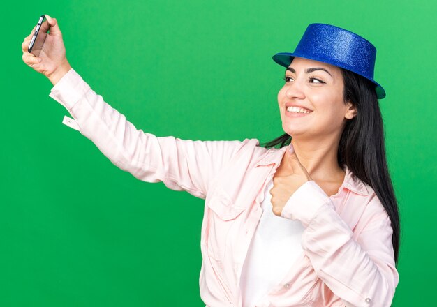 Smiling young beautiful girl wearing party hat take a selfie showing thumb up