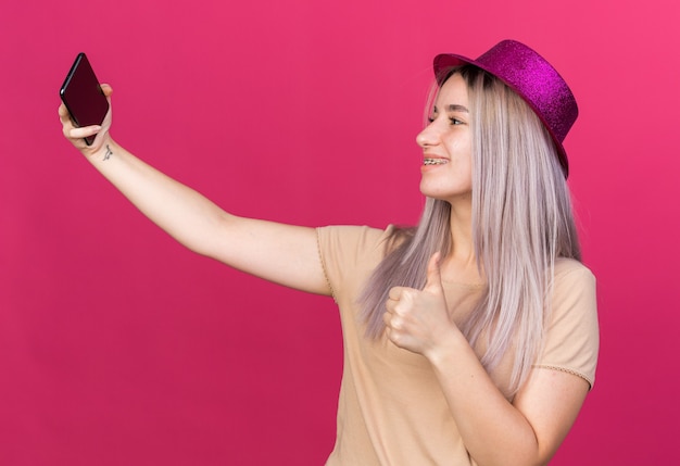 Smiling young beautiful girl wearing party hat take a selfie showing thumb up 