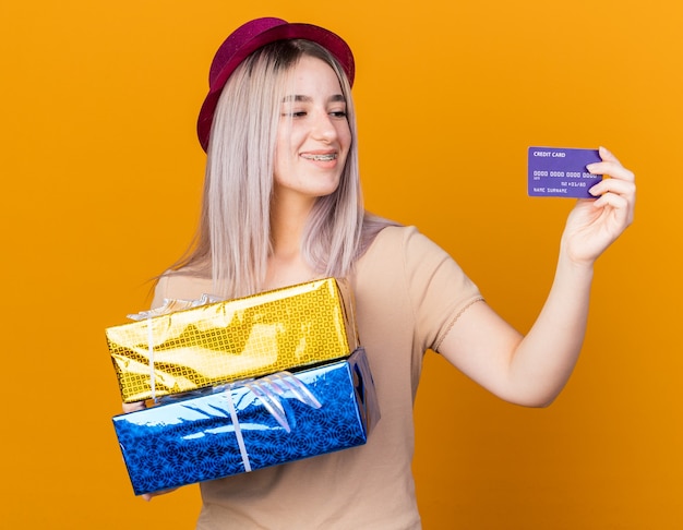 Бесплатное фото Улыбающаяся молодая красивая девушка в партийной шляпе с подтяжками держит подарочные коробки и смотрит на кредитную карту в руке, изолированной на оранжевой стене