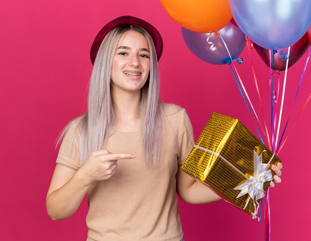 Улыбающаяся молодая красивая девушка в шляпе с зубными скобами, держащая воздушные шары с подарочной коробкой