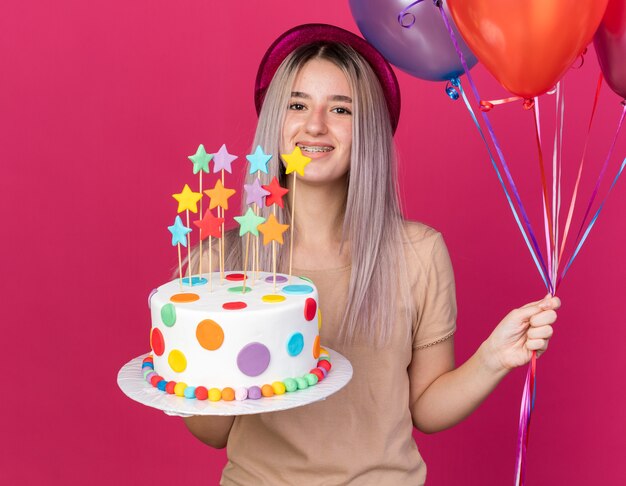 Улыбающаяся молодая красивая девушка в шляпе с зубными скобами, держащая воздушные шары с тортом
