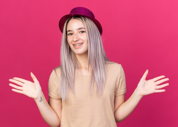 Smiling young beautiful girl wearing party hat with dental braces spreading hands 