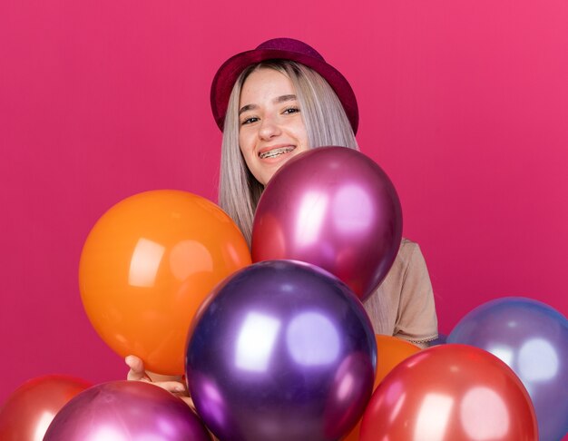 Smiling young beautiful girl wearing party hat with dental braces standing behind balloons 