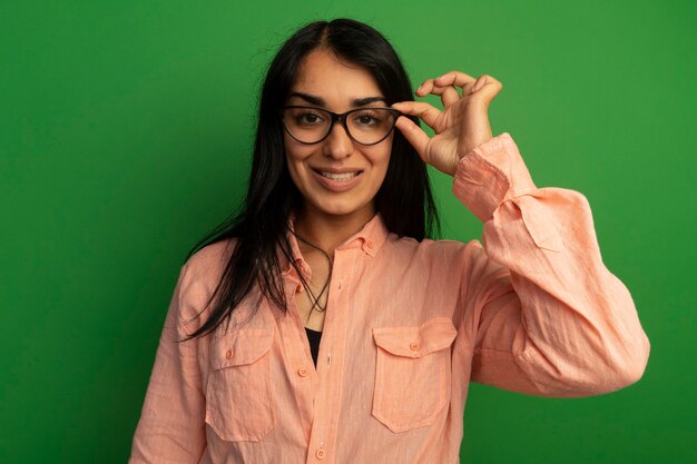 분홍색 티셔츠를 입고 녹색 벽에 고립 된 안경을 들고 웃는 젊은 아름다운 소녀