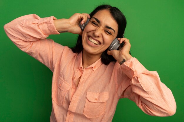 Smiling young beautiful girl wearing pink t-shirt with headphones isolated on green wall
