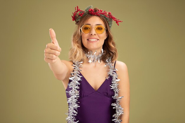 紫色のドレスとメガネを身に着けている笑顔の若い美しい少女の首に花輪と花輪がオリーブグリーンの背景で隔離の親指を示しています