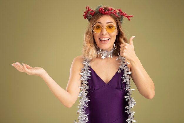 Free photo smiling young beautiful girl wearing purple dress and glasses with wreath and garland on neck showing thumb up isolated on olive green background