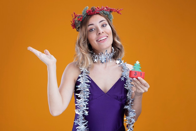 Smiling young beautiful girl wearing purple dress and wreath with garland on neck holding christmas toy points with hand at side isolated on brown background with copy space