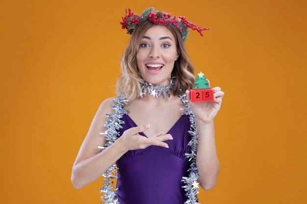 紫色のドレスと花輪を身に着けている若い美しい少女の笑顔と茶色の壁に分離されたクリスマスのおもちゃを指しています。