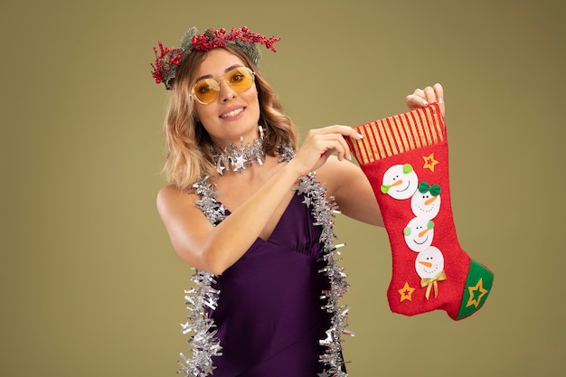 올리브 녹색 배경에 고립 된 크리스마스 양말을 들고 목에 보라색 드레스와 안경 및 화 환을 입고 젊은 아름 다운 소녀 미소