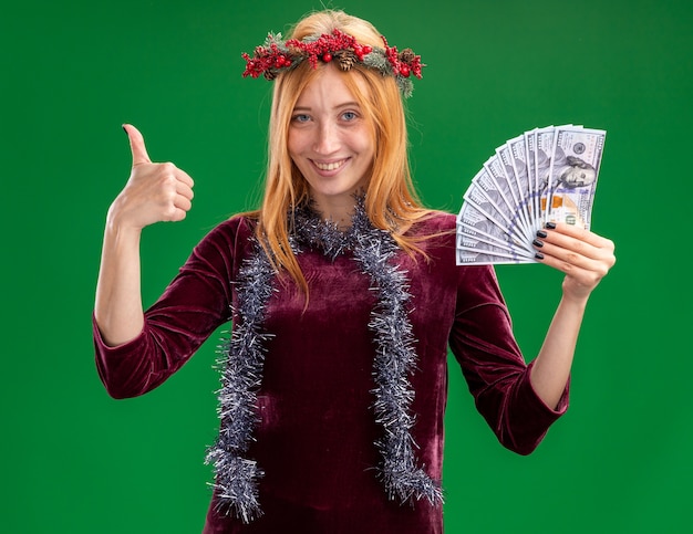 smiling young beautiful girl wearing red dress with wreath and garland on neck holding cash showing thumb up isolated on green wall