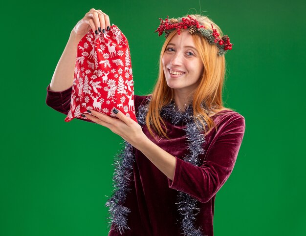 Smiling young beautiful girl wearing red dress with wreath and garland on neck holding christmas bag isolated on green background