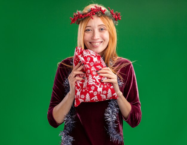 녹색 배경에 고립 된 크리스마스 가방을 들고 목에 화 환과 갈 랜드와 빨간 드레스를 입고 웃는 젊은 아름 다운 소녀