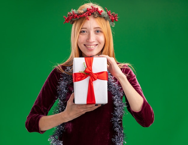 Smiling young beautiful girl wearing red dress with wreath and garland on neck holding gift box isolated on green background