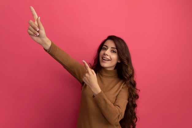 Smiling young beautiful girl wearing turtleneck sweater points at side isolated on pink wall