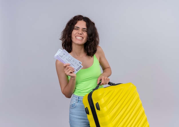 Smiling young beautiful traveler woman holding airplane tickets and suitcase on isolated white wall with copy space