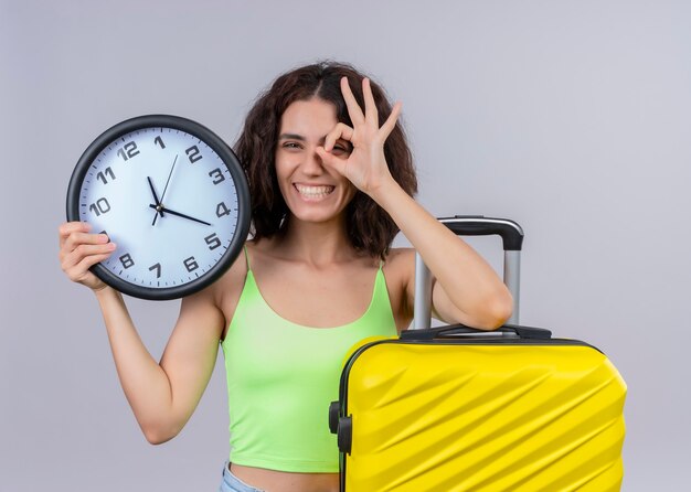Smiling young beautiful traveler woman holding clock and suitcase and doing look gesture on isolated white wall
