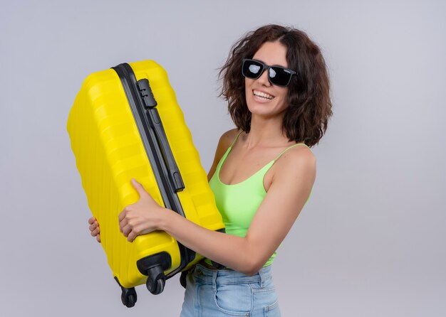 Smiling young beautiful traveler woman wearing sunglasses and holding suitcase on isolated white wall