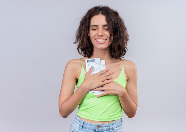 Smiling young beautiful woman holding airplane tickets with closed eyes on isolated white wall with copy space