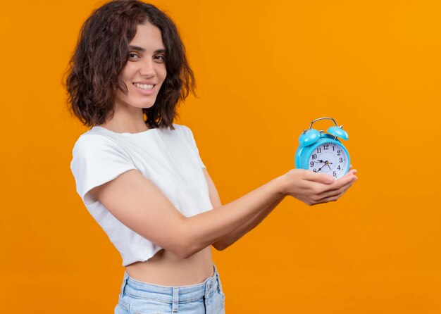 Smiling young beautiful woman holding alarm clock on isolated orange wall