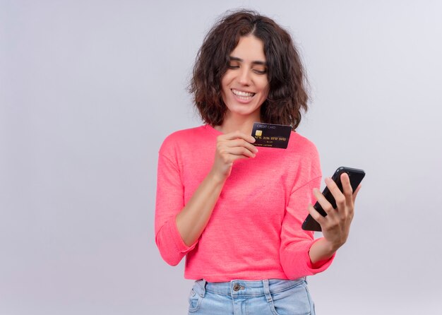 Smiling young beautiful woman holding card and mobile phone on isolated white wall with copy space